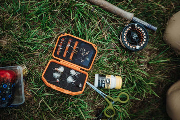 Orange Atollas Micro Fly Box opened up with flies displayed on the grass floor next to a fly rod.