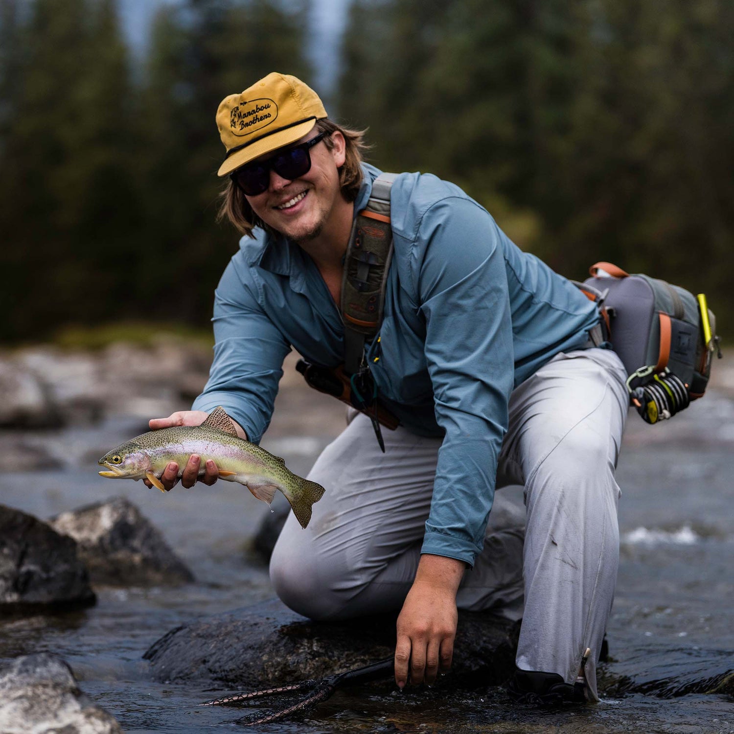 A smiling fly fisherman kneels in a river holding a rainbow trout, wearing a yellow cap with a visible 'Marabou Brothers' logo, a blue long-sleeve shirt, light gray pants, and a fishing backpack.