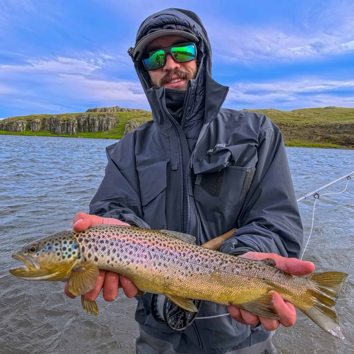 A fly fisherman wearing a black hooded waterproof jacket and mirrored sunglasses stands in shallow water holding a brown trout fly fishing rod in the background of a lake with rocky cliffs under a blue sky.