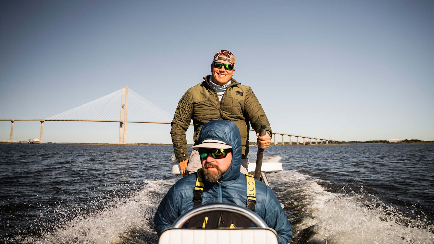 Two fly fisherman on a skiff during the day speeding down a waterway with a bridge in the background during the day