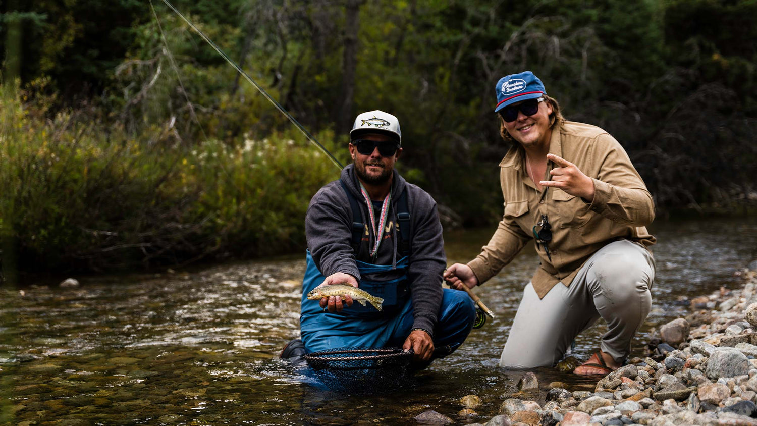 Two fly fishermen kneeling in a trout stream holding a trout with green forest in background