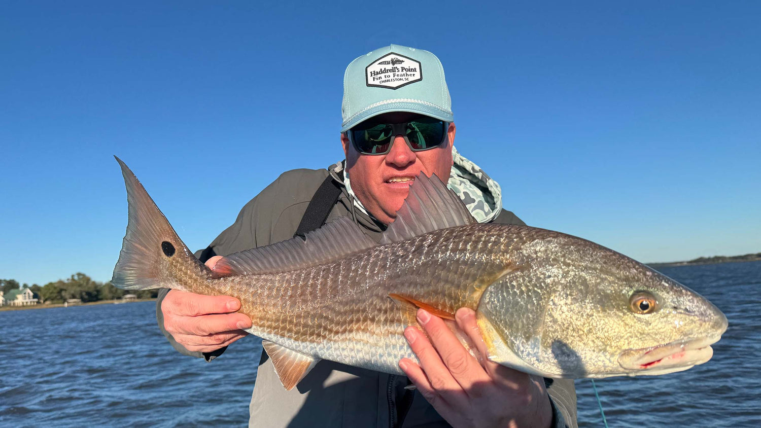 Atollas Ambassador Adam Motes wearing a light blue 'Haddrell's Point Fin to Feather' cap, sunglasses, and a gray hoodie holds a large redfish on a fishing line over a body of water under a clear blue sky.