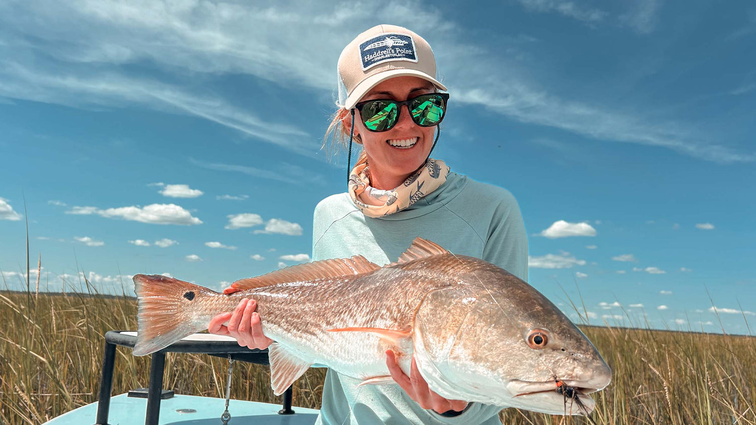 A smiling female fly angler with a tan hat and sunglasses holding a large redfish she caught in the marsh flats with blue sky in the background.