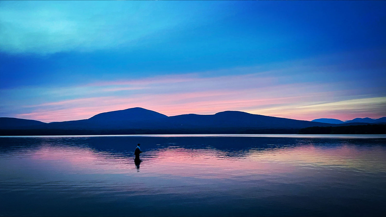 A scenic landscape photograph of a calm lake at dusk or dawn, with layered blue mountains in the distance and a small silhouetted figure standing in the water near the center. The sky transitions from deep blue to soft pink and purple, reflected clearly on the water surface.