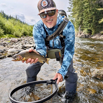 A smiling fly angler in a blue camouflage fly-landing shirt, waders, and cap stands in a rocky river holding a trout, with a net in the foreground and forested banks in the background.
