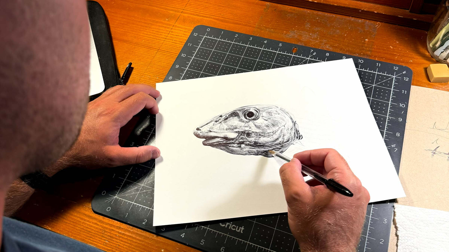 An artist drawing the head of a bonefish on a white piece of paper on wooden table indoors.