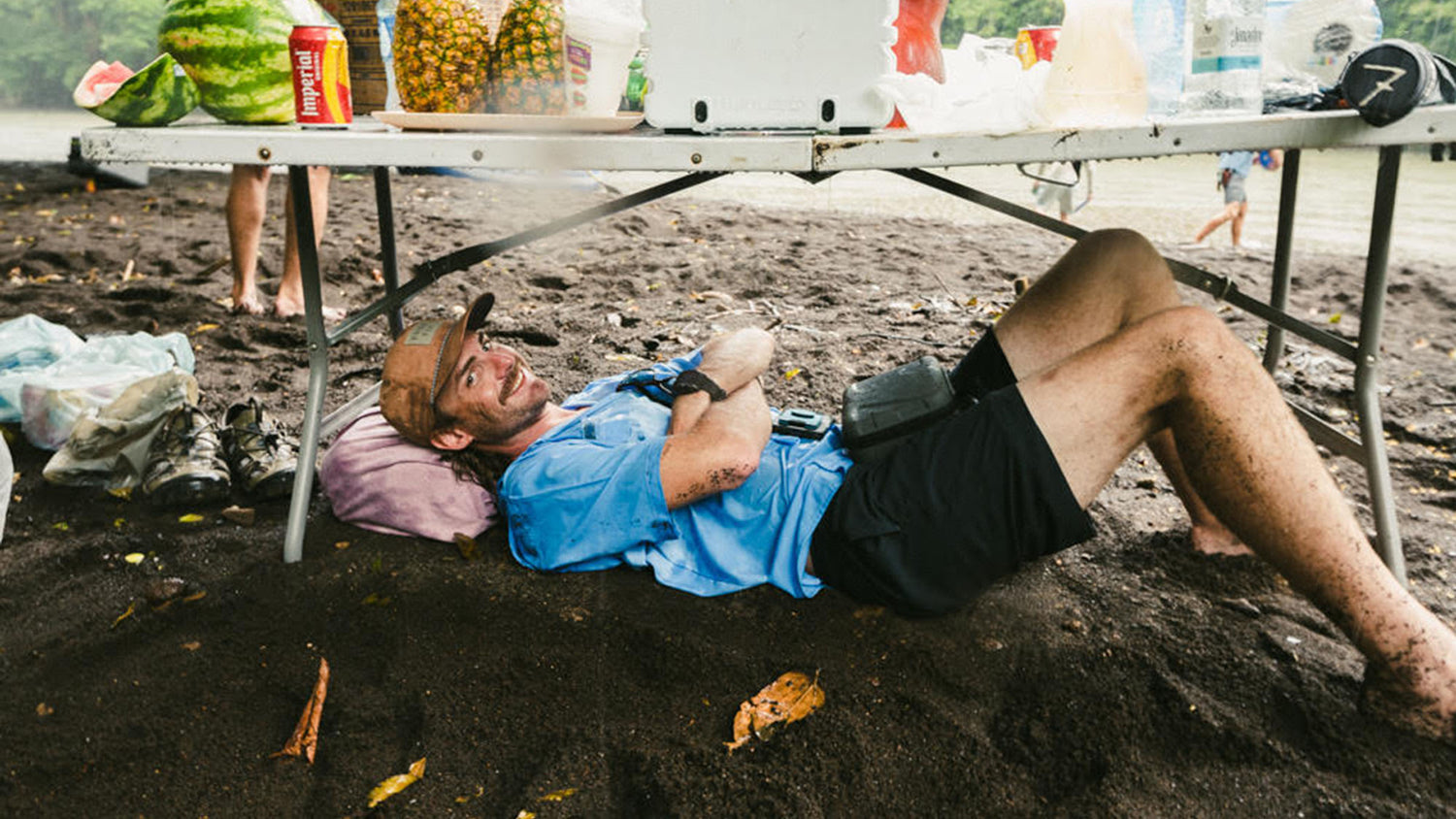 Atollas Ambassador Charles Hill lies on dark sand under a folding table on a beach, wearing a light blue polo shirt, black shorts, and a tan cap. A cooler, watermelons, and canned drinks are visible on the table, with water and shoreline in the background.