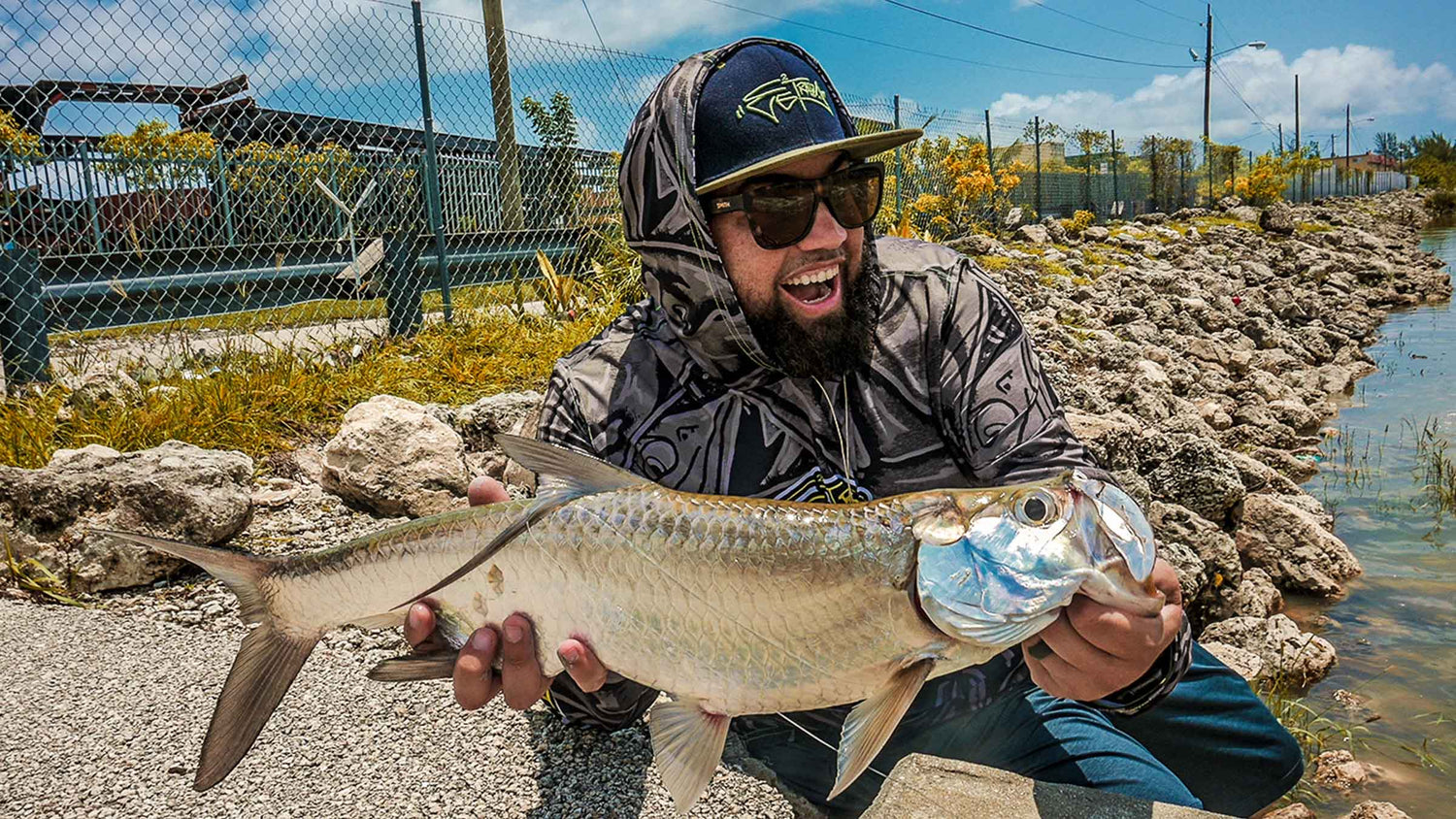 A fly fisherman wearing a black and gray patterned hoodie, sunglasses, and a cap holds a large silver tarpon in an outdoor rocky area next to water.