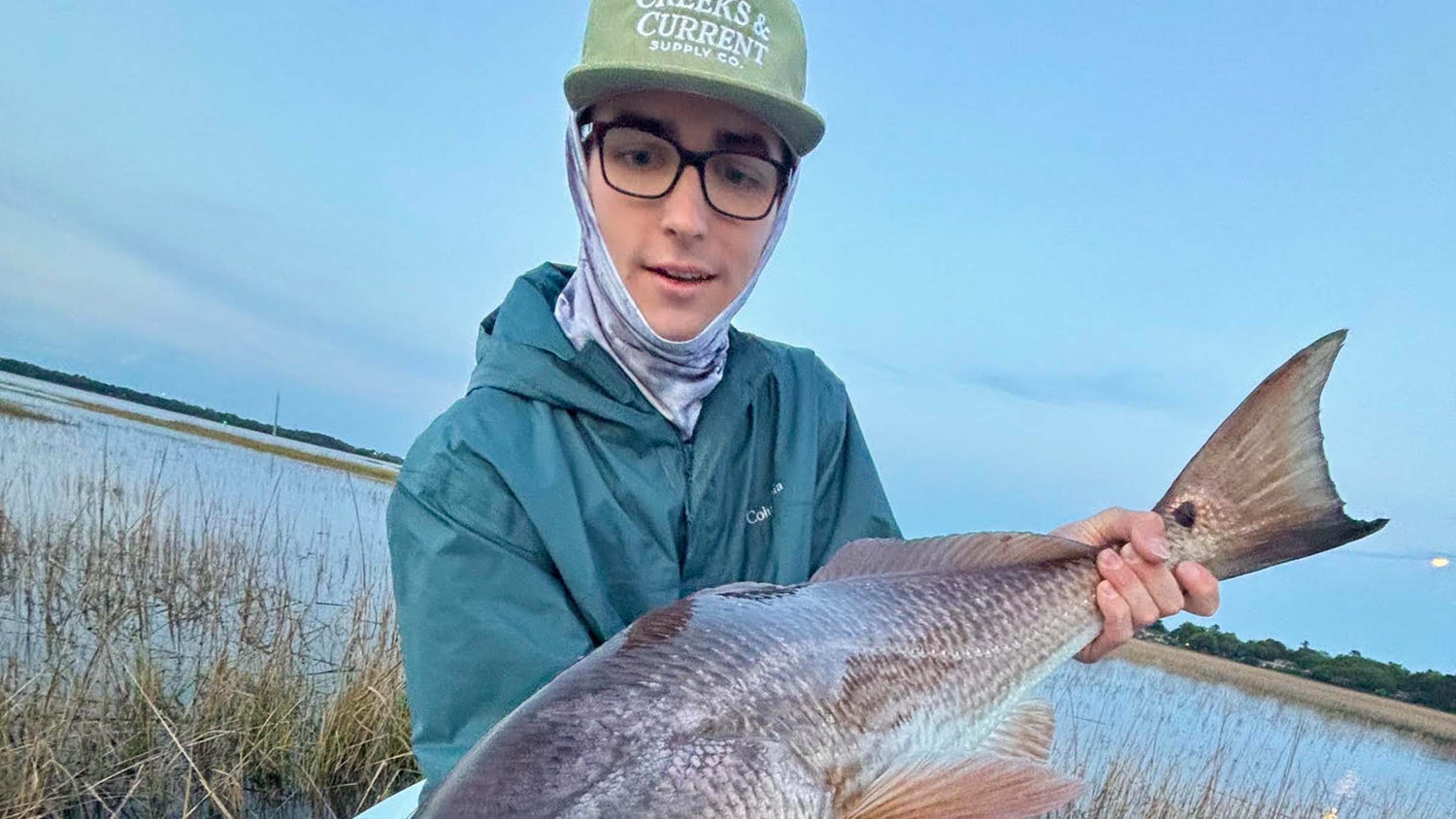 A fly fisherman wearing a green cap, glasses, a face covering, and a teal hooded jacket holds a large redfish in a marsh under a blue sky.
