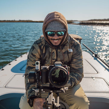 A male fly fisherman with a camo jacket, hoodie, and sunglasses on a skiff with a professional camera pointing at the viewer in the marsh creeks during the day.
