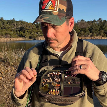 A fly fisherman outdoors by a lake wearing a camouflage cap with a 'Drum Spots' logo, a green fleece jacket, and an olive green Atollas 1GO Fly Box across his chest while examining a fishing fly.