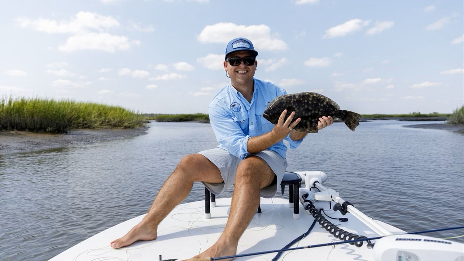 A fly fisherman wearing a light blue shirt, gray shorts, and a blue cap sits on a white boat holding a large flounder in a river channel with marsh grass on the side under a partly cloudy sky.