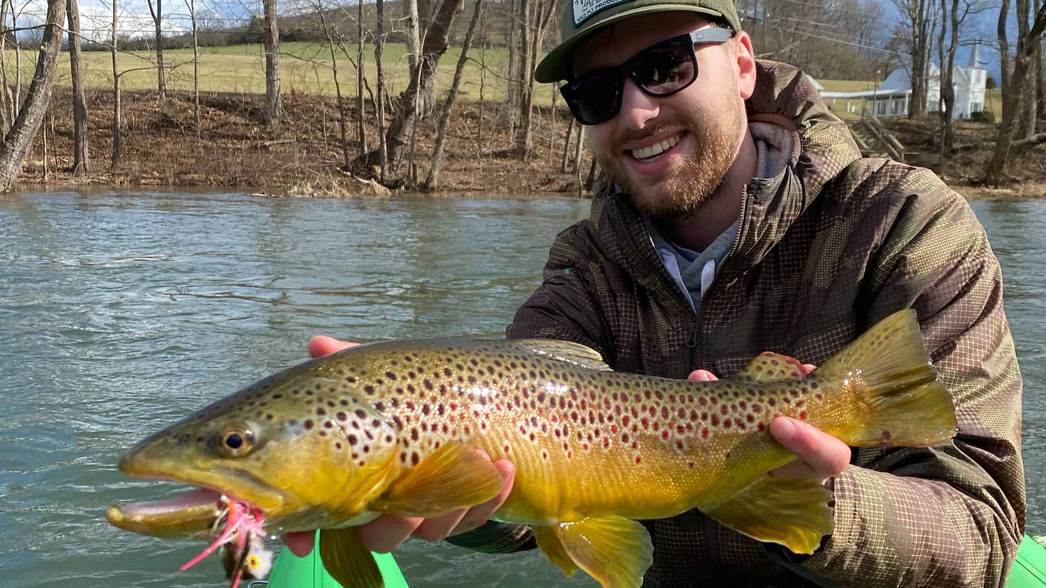 A smiling fly fisherman wearing sunglasses and a cap holds a large brown trout fish with a pink fly in its mouth, with a river and wooded shoreline in the background.