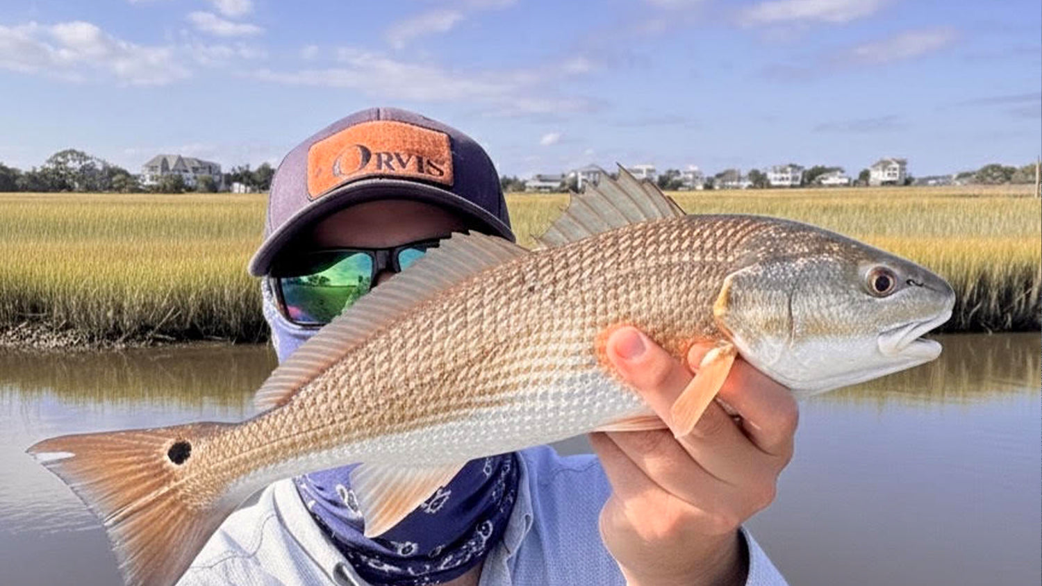 A fly fisherman outdoors by the water holding a redfish, wearing a dark cap with an orange patch that reads 'ORVIS' and sunglasses, with marsh grass and houses in the background.
