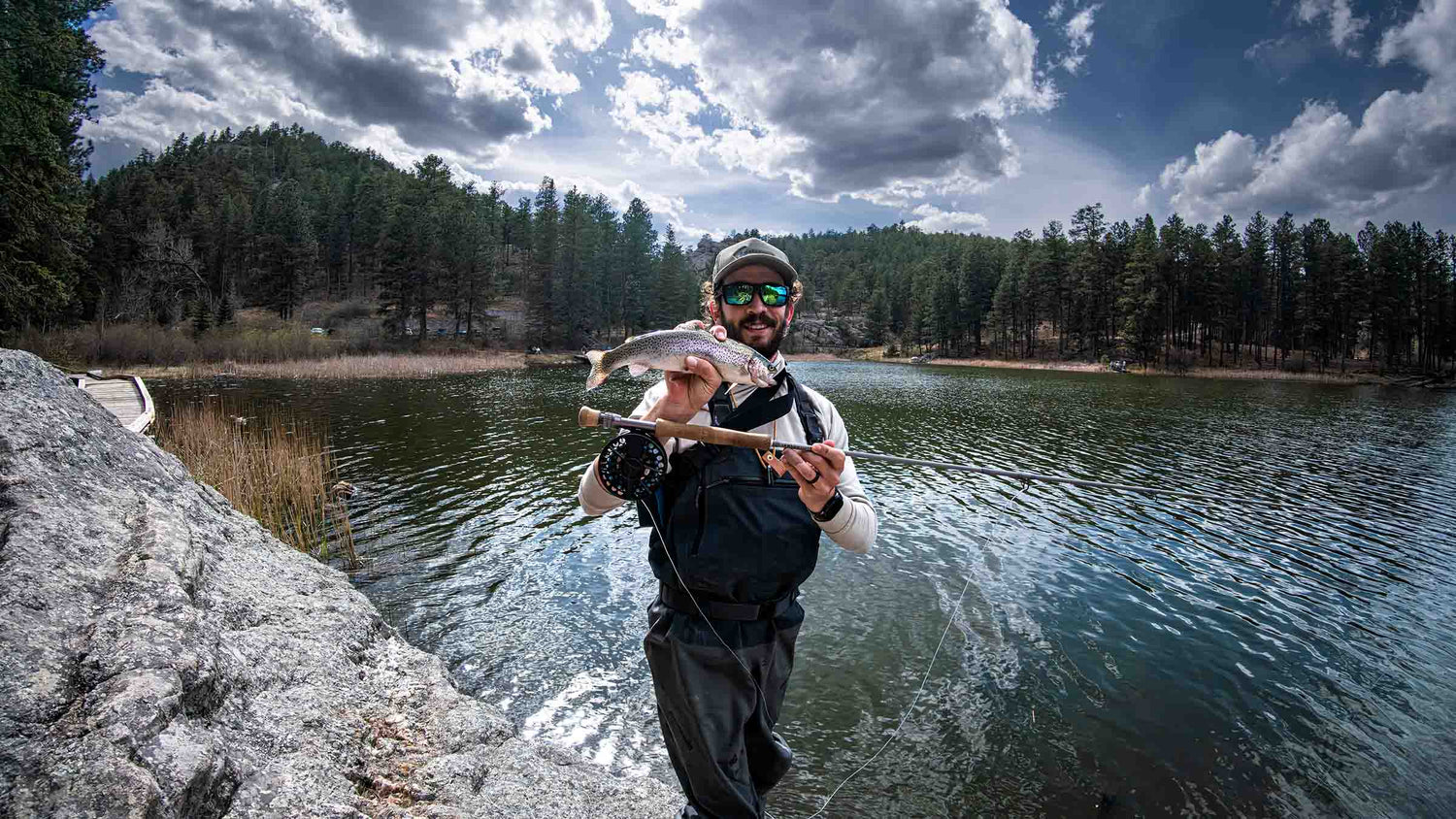 A fly angler wearing a black fishing vest and sunglasses stands on the edge of a lake, holding a freshly caught fish over his shoulder, with a forested shoreline and dramatic clouds in the background.