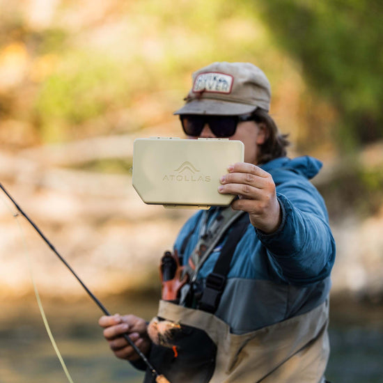 Fly angler holding a sand colored Atollas 2GO Fly Box showing the backside of it with the Atollas logo showing against a blurry outdoor background