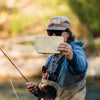 Fly angler holding a sand colored Atollas 2GO Fly Box showing the backside of it with the Atollas logo showing against a blurry outdoor background