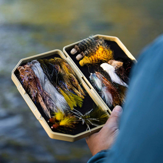 Fly angler holding a sand colored Atollas 2GO Fly Box with assorted colorful flies inside against a blurred watery background