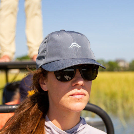 Fly angler wearing a slate blue cap with an Atollas logo and sunglasses, sitting in a boat with a blurred natural background