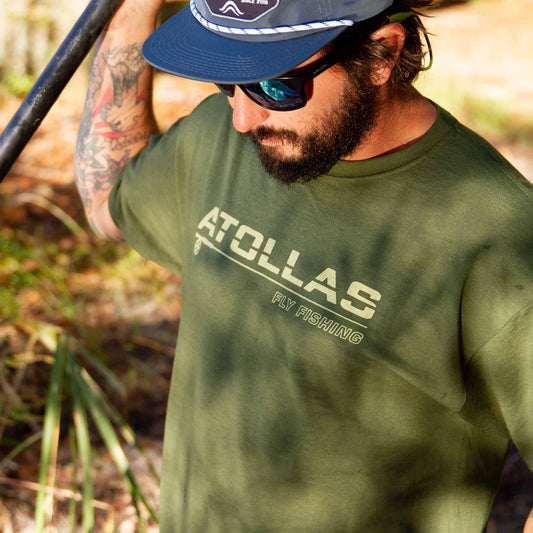 Fly angler wearing a green t-shirt with 'ATOLLAS' fly fishing branding outdoors.
