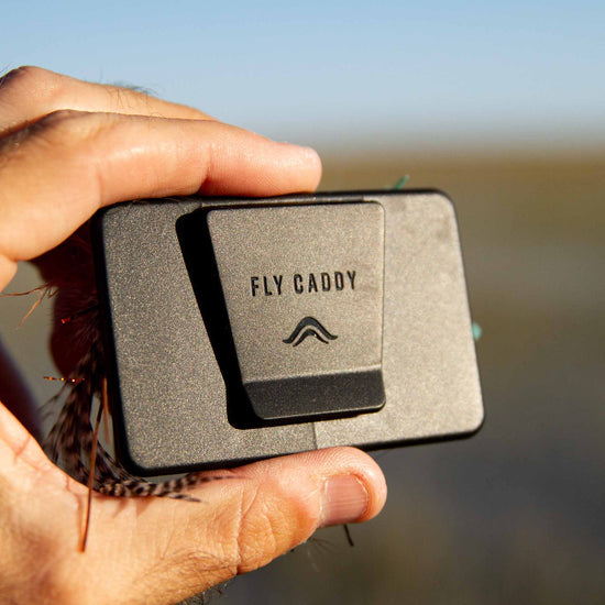 A hand holding an Atollas Fly Caddy fly patch showing the backside which has a clip on it against a blurry background