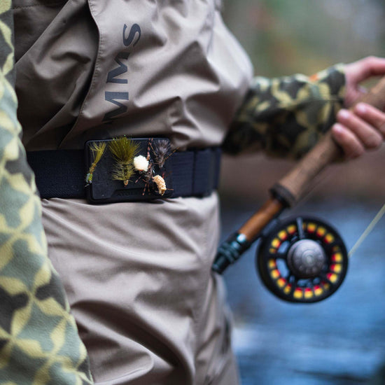 A person holding a fly rod with an Atollas Fly Caddy fly patch clipped to their wader belt with a blurry forested background