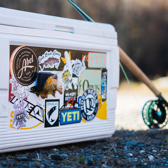 White cooler with an assortment of fly fishing stickers on it along with an Atollas Fly Caddy fly patch clipped into a docking station that is adhered to the cooler