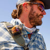 Man wearing sunglasses and a plaid shirt with a backpack against a clear blue sky and Fly Caddy fly patch clipped to his shoulder strap