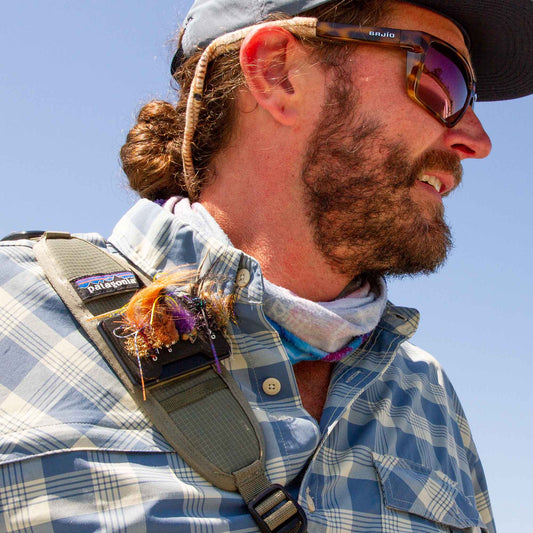 Man wearing sunglasses and a plaid shirt with a backpack against a clear blue sky and Fly Caddy fly patch clipped to his shoulder strap