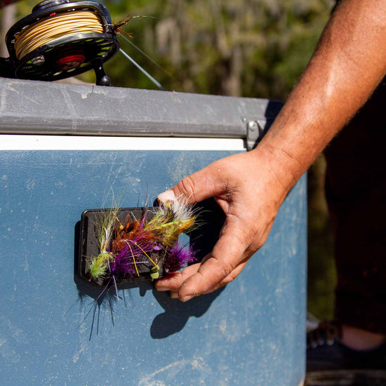 A hand reaching for an Atollas Fly Caddy XL fly patch which is clipped into a docking station attached to a blue cooler with a fly rod on top of it