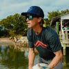 Fly angler wearing a dark colored Atollas logo shirt and sunglasses by a body of water with trees in the background