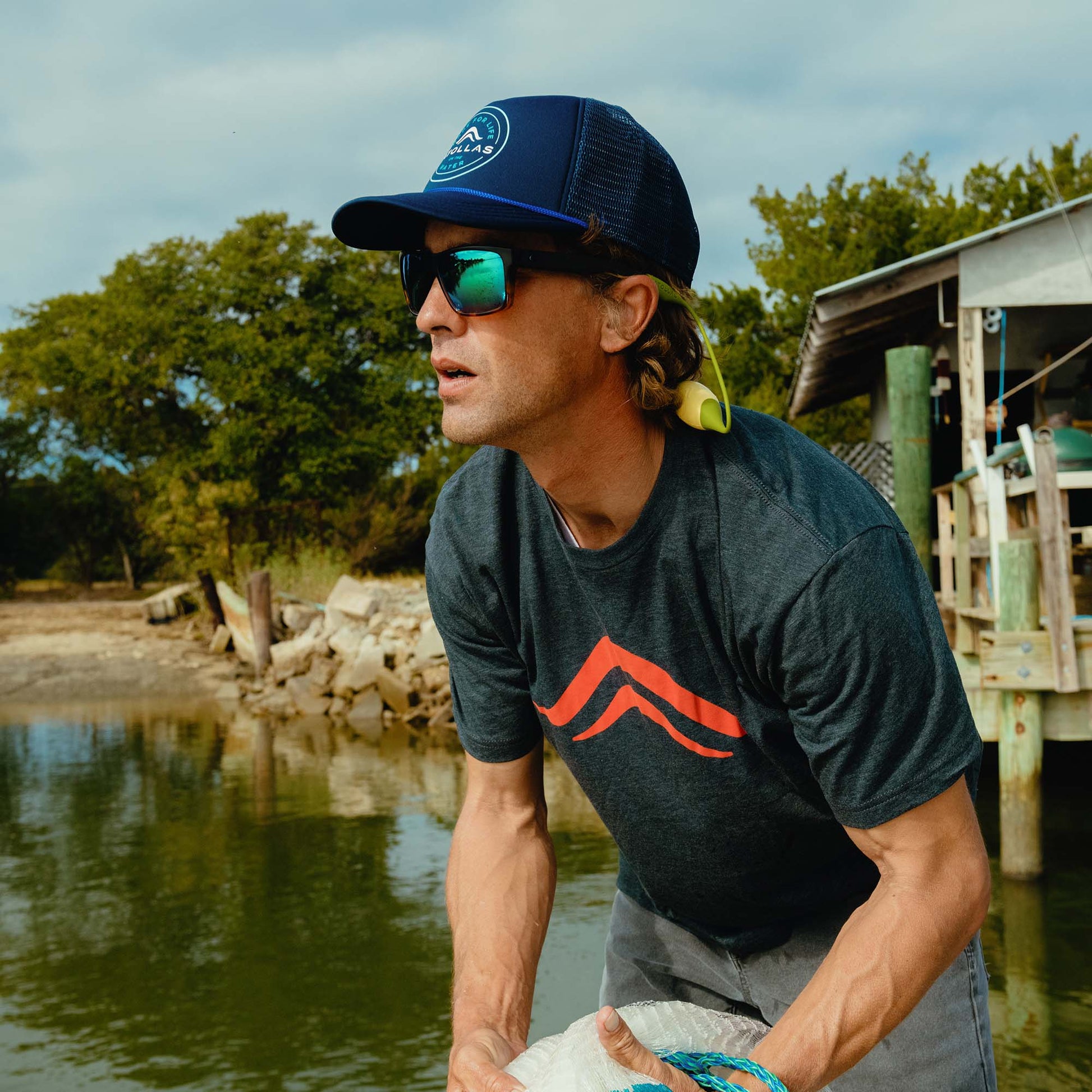 Fly angler wearing a dark colored Atollas logo shirt and sunglasses by a body of water with trees in the background
