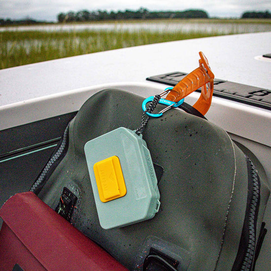 Green Atollas Micro Fly Box attached to a waterproof pack with a carabiner, with a blurred outdoor background.
