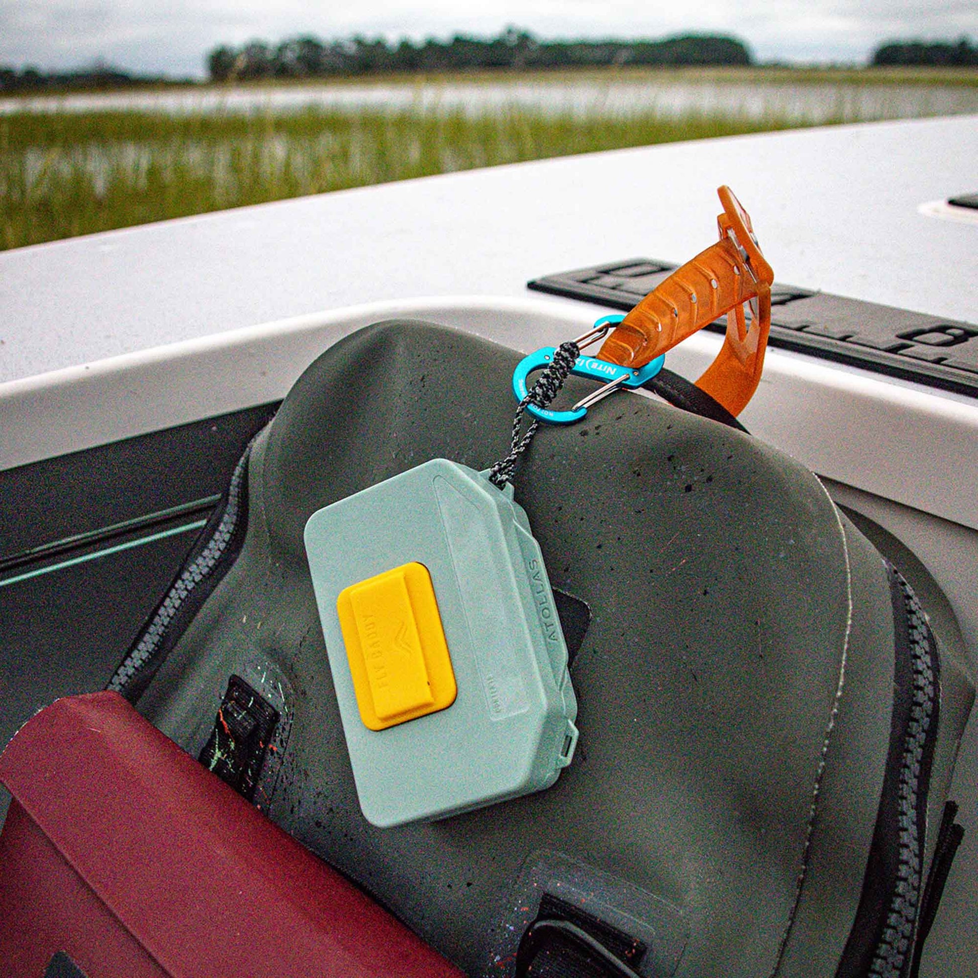 Green Atollas Micro Fly Box attached to a waterproof pack with a carabiner, with a blurred outdoor background.