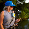 Fly angler fishing by a river, wearing a blue cap and sunglasses, with trees in the background and Atollas Micro Fly Box attached to the sling of their pack on their shoulder