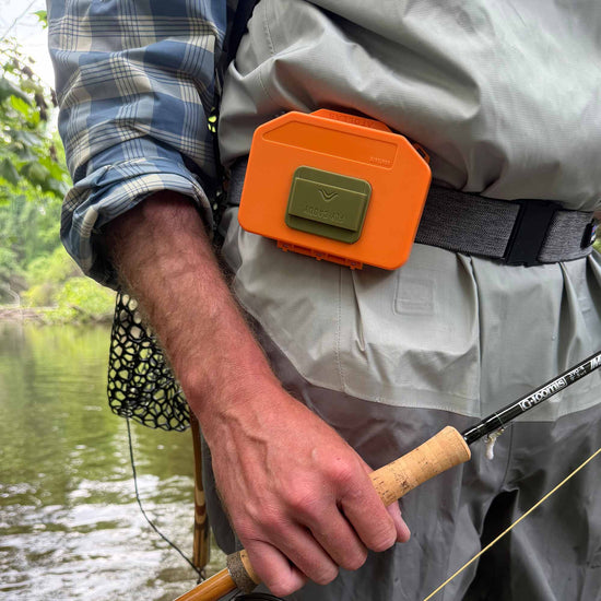 Fly angler holding a fishing rod with an orange Atollas Micro Fly Box clipped on their wader belt, standing by a body of water
