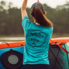 Fly angler wearing a teal shirt with 'Atollas' logo, standing next to a kayak.