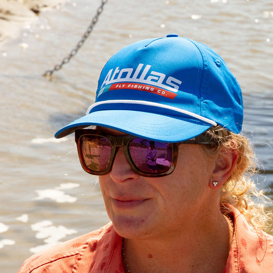 Fly angler wearing a blue cap with 'Atollas Fly Fishing Co' logo and sunglasses, standing by water.