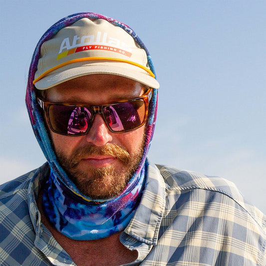 Fly angler wearing a colorful headscarf, sunglasses, and an Atollas fly fishing co. cap against a clear sky.